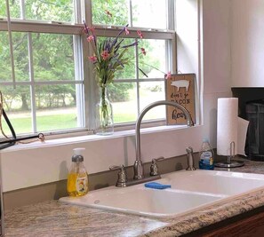 A kitchen with a white sink, a window with a view of greenery, a soap dispenser, and a paper towel holder. - Wildflower Cottage : Country Home Near Town (Paragould)