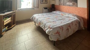 Une chambre avec un plafond en bois, un lit recouvert d’une literie à motifs floraux, un téléviseur posé sur un meuble en bois et une fenêtre avec des rideaux.
