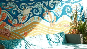 A room with a bed, a sofa, and a wall tapestry depicting a tropical scene. - Lisbon Soul Surf Camp (Cascais)