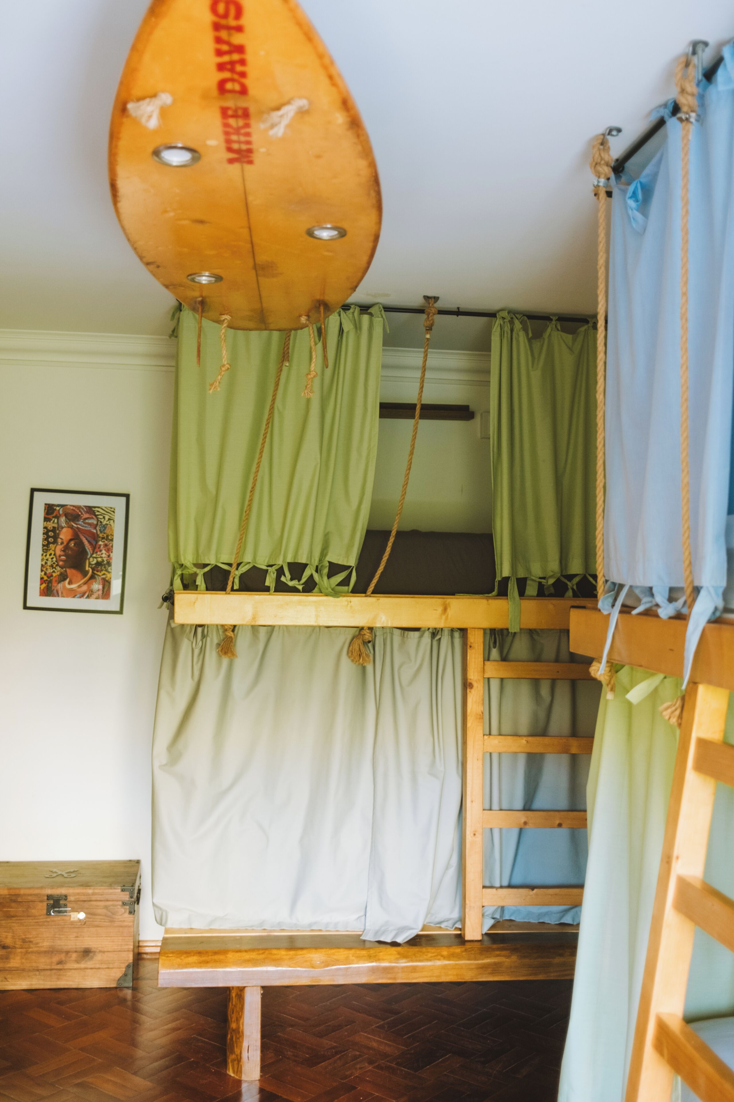 A bunk bed with green curtains, a wooden bench, and a surfboard with 'MIKE DAYTON' written on it.