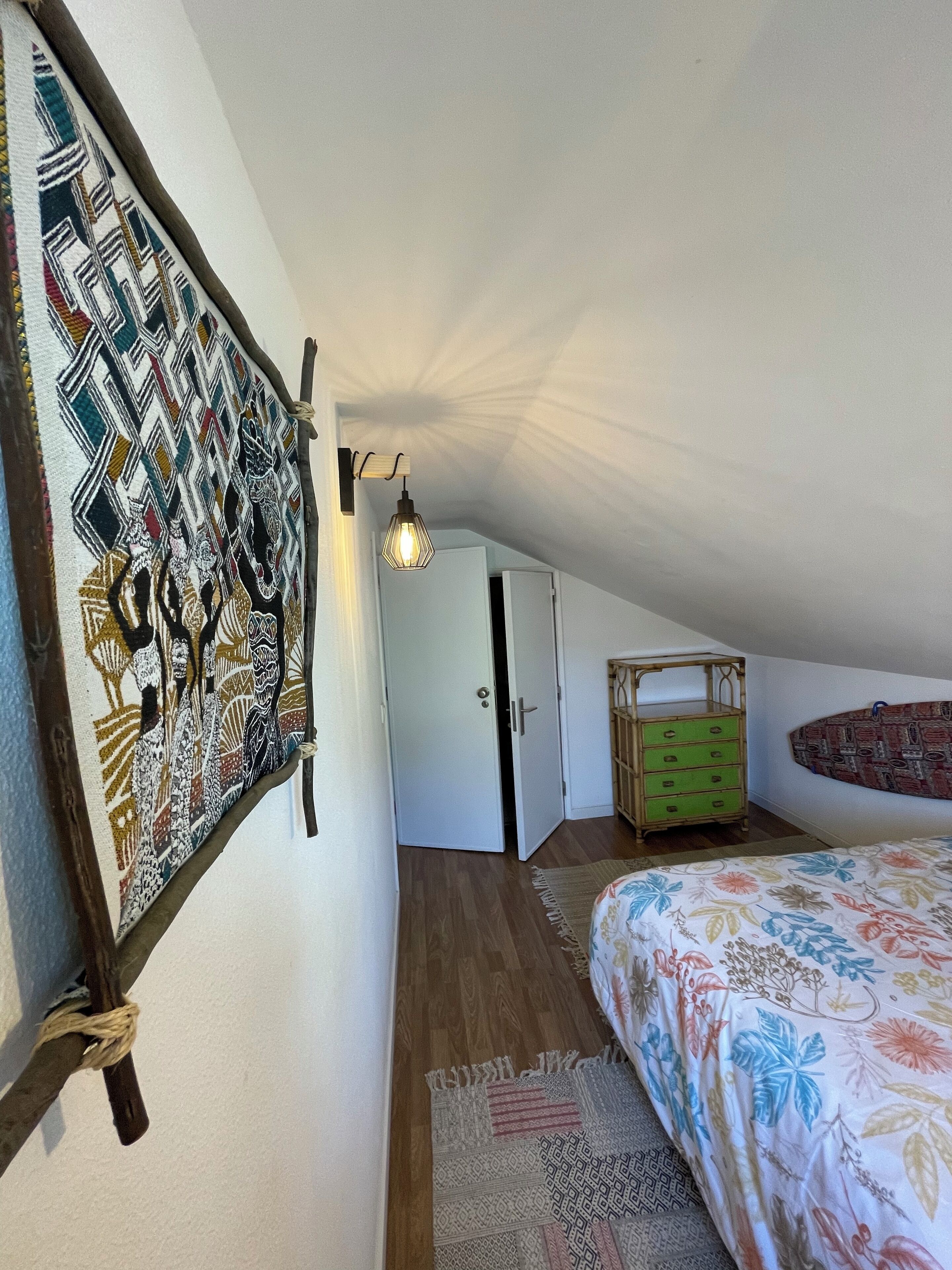 A bedroom with a sloped ceiling, a bed with a floral bedspread, a wooden dresser, a wall-mounted shelf, and a hanging light fixture.