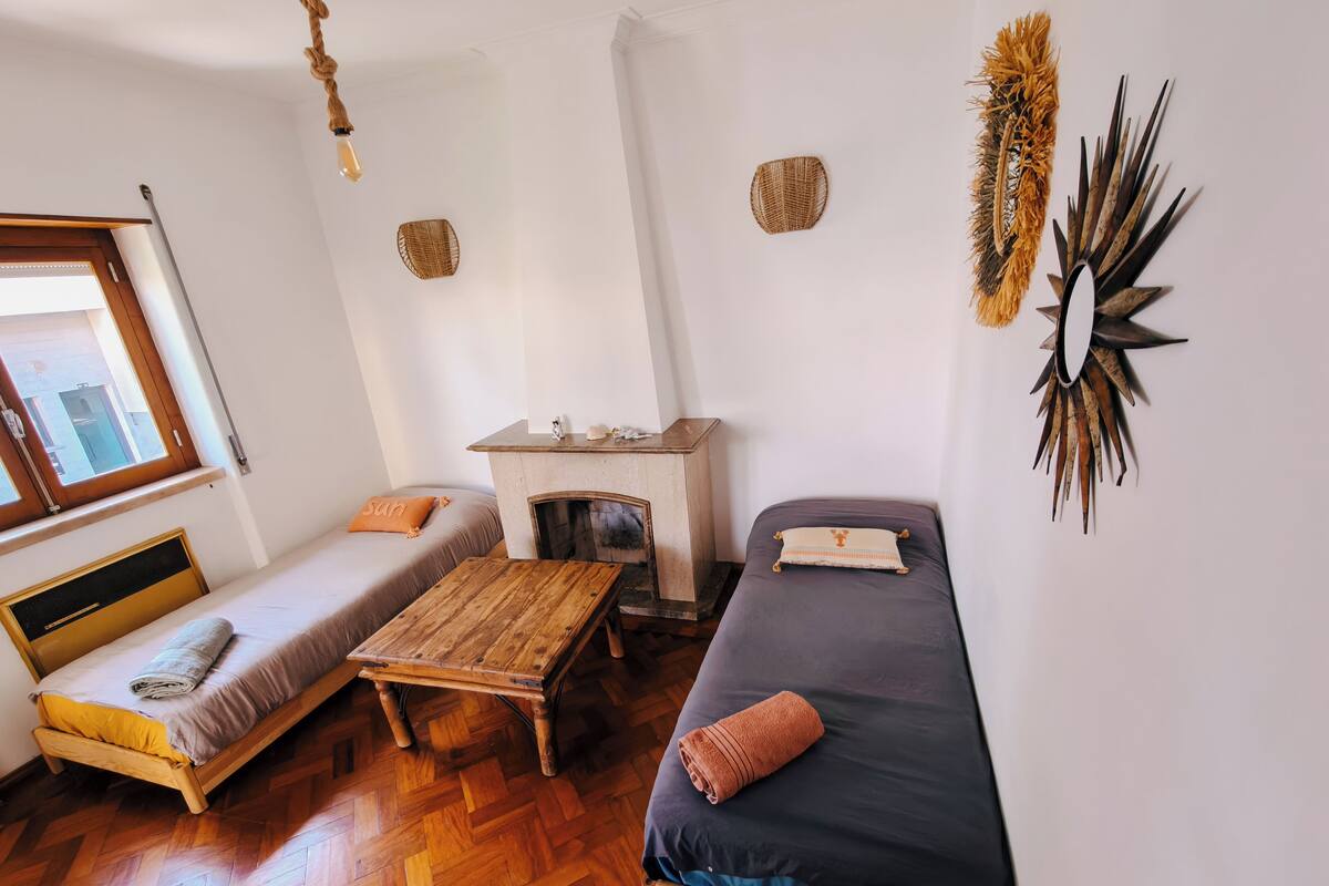 A room with two beds, a wooden table, a fireplace, and a hanging light fixture.