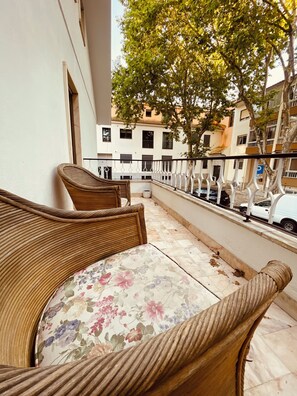 Un balcon agrémenté de meubles en osier et d’une nappe à motifs floraux, donnant sur une rue bordée d’immeubles et de voitures garées.