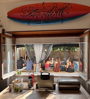 Un coin repas décoré avec une planche de surf, une table avec une machine à café et une vue sur les personnes à l’extérieur.