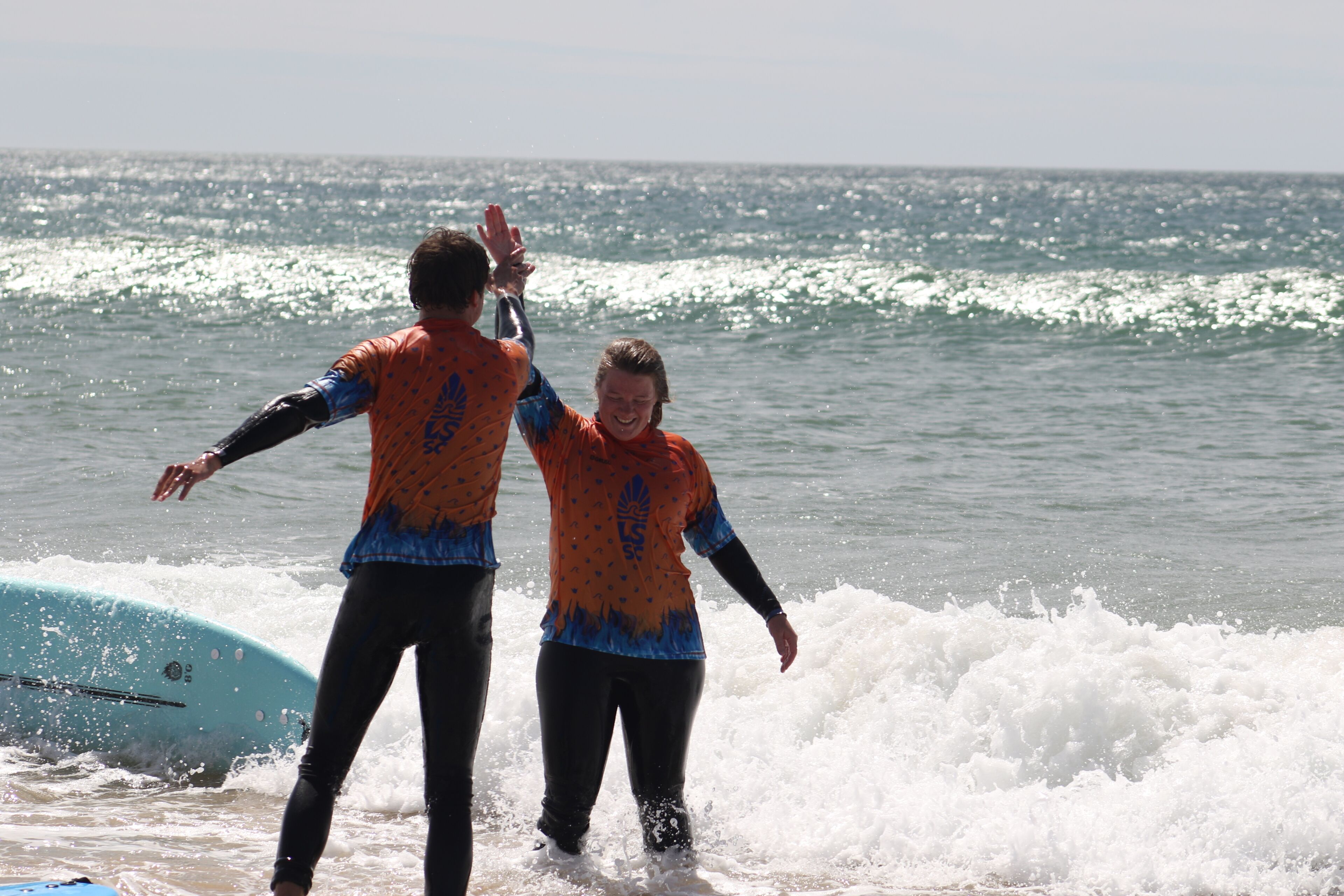 Deux personnes en combinaisons de plongée, debout dans une eau peu profonde, avec une planche de surf.