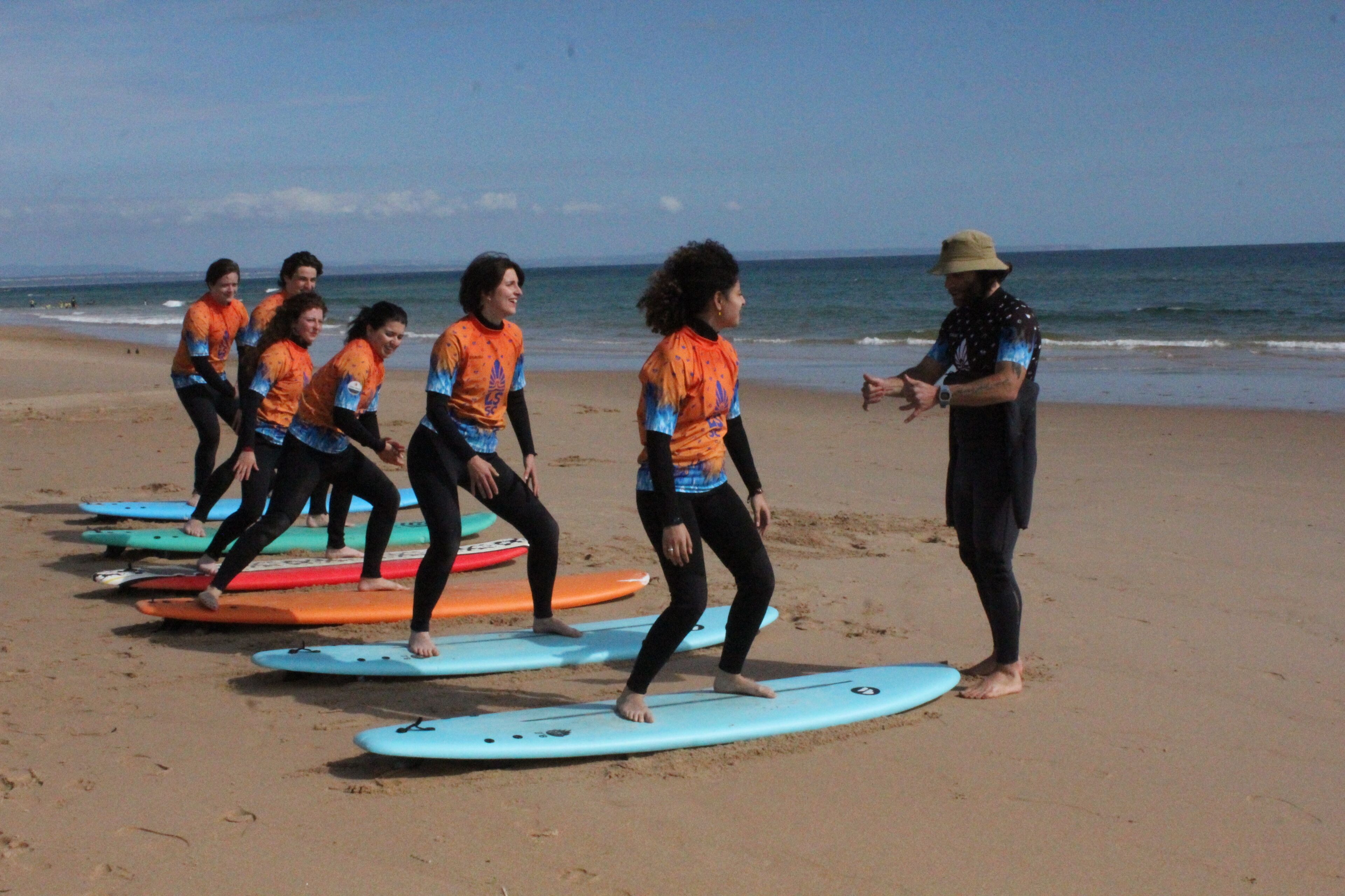 Een groep mensen op het strand met surfplanken, een persoon met een hoed, en de oceaan op de achtergrond.