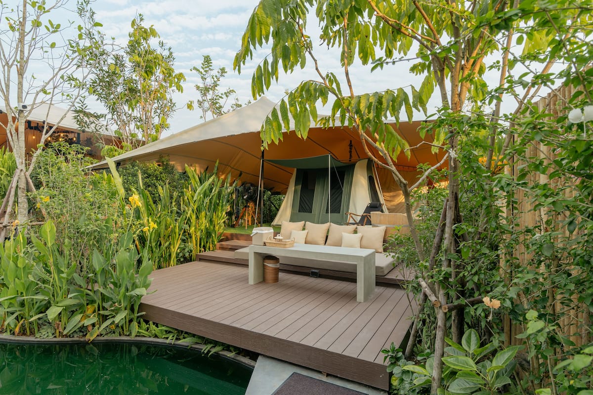 Uma área de estar moderna ao ar livre, com banco e almofadas, cercada por vegetação exuberante e uma estrutura de tenda.