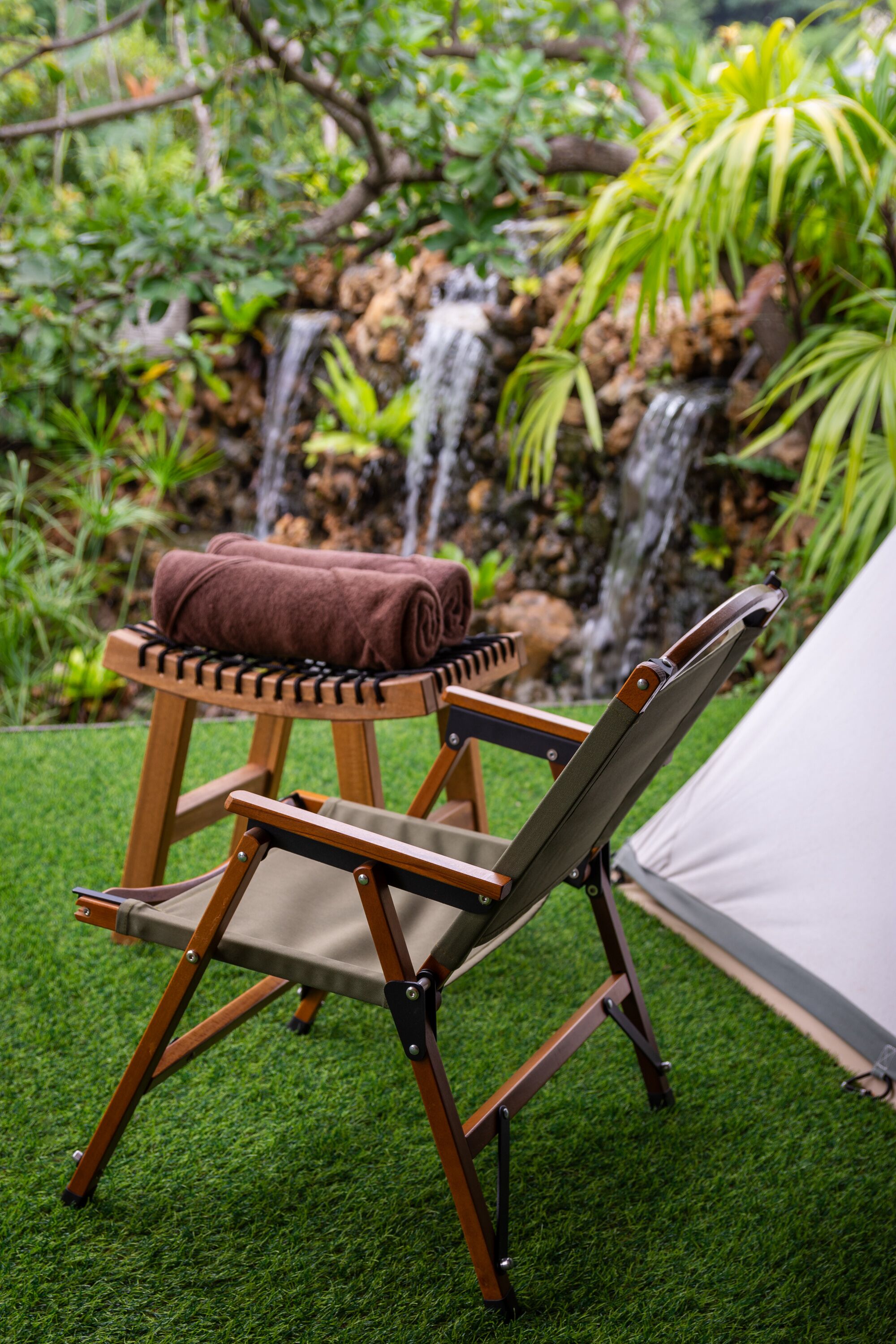 Een houten stoel met een handdoek, een klein tafeltje en een tent in een tuin met een waterval.
