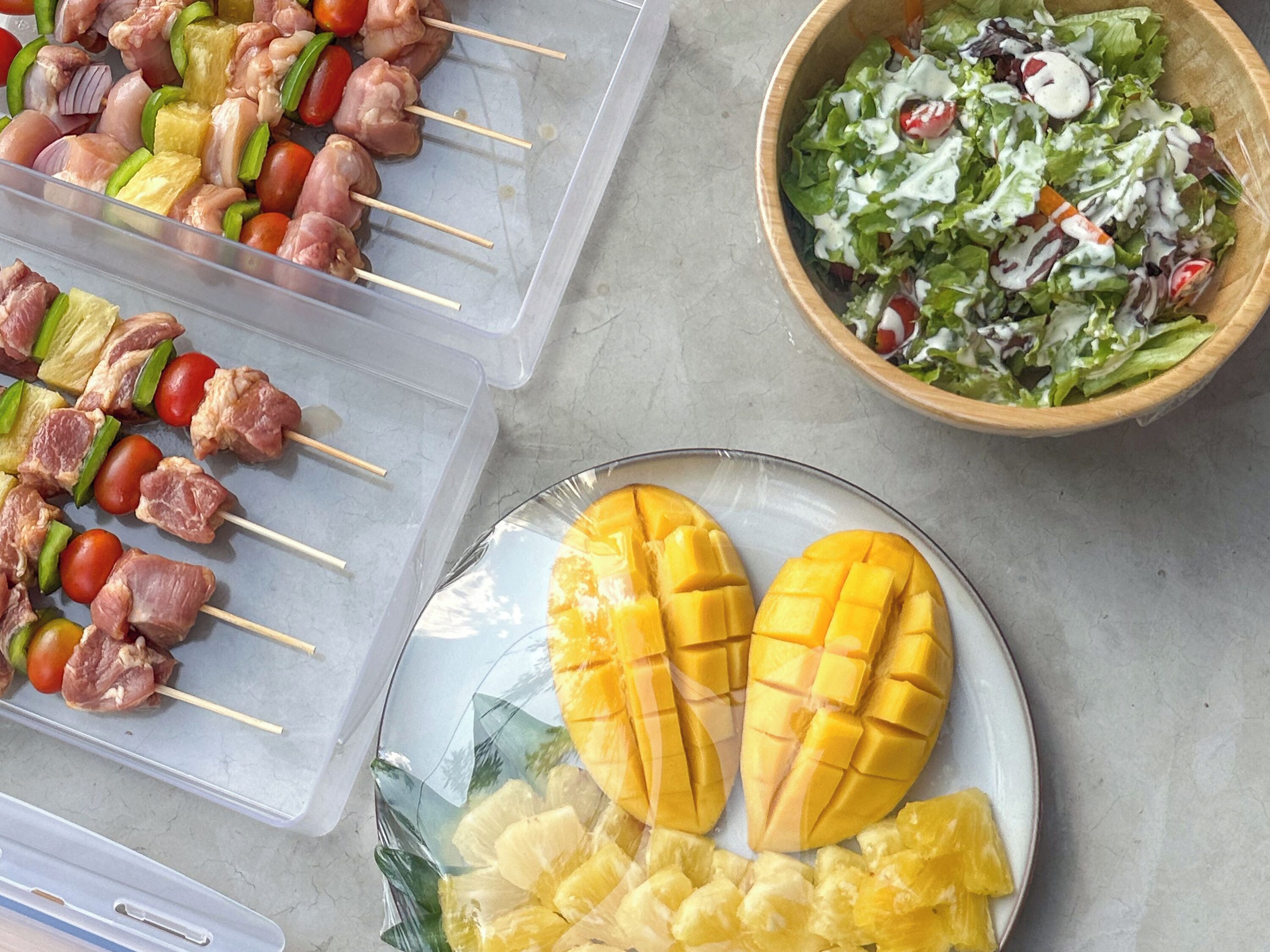 A table with skewers of meat and vegetables, a bowl of salad, and a plate of mango and pineapple.