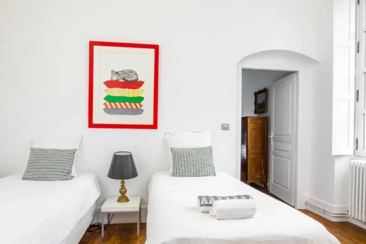 A hotel room with two beds, a framed picture on the wall, a lamp, and a door leading to another room.