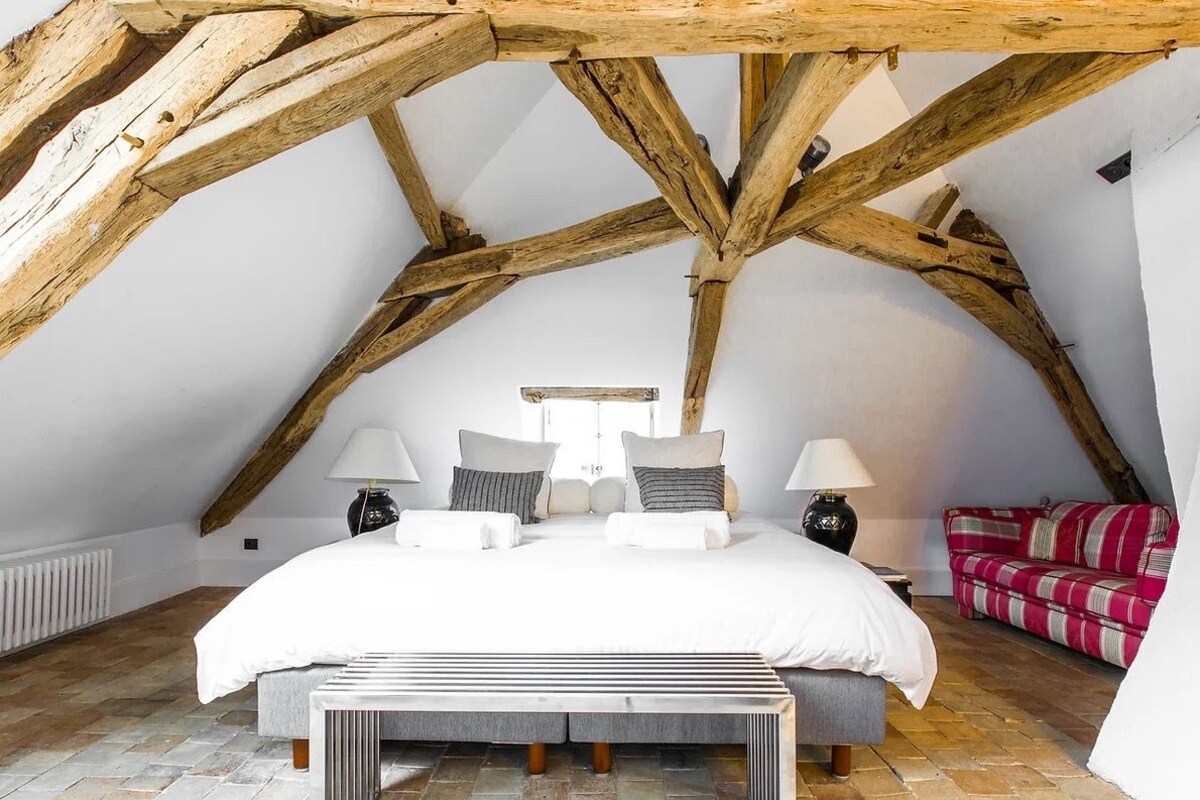 A bedroom with a sloped ceiling, a bed with white bedding, two bedside tables with lamps, a red and white striped sofa, and a stone-patterned floor.