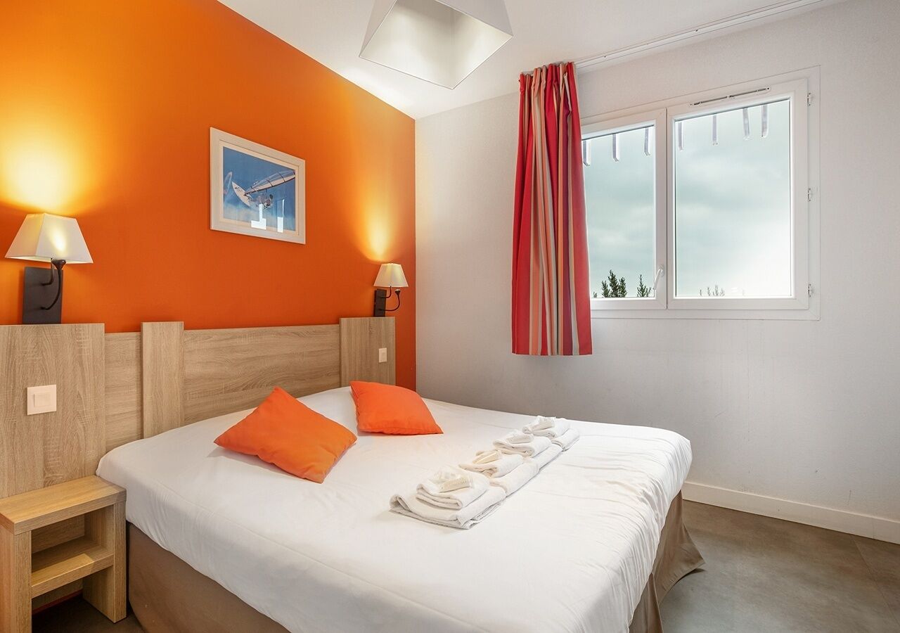 A hotel room with a bed, two pillows, a nightstand, a lamp, a window with curtains, and a picture on the wall.