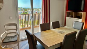 Ein Zimmer mit einem Holzesstisch, sechs Stühlen, einem Fernseher auf einer Holzkommode und einem Balkon mit Blick auf Gebäude und Grünflächen.