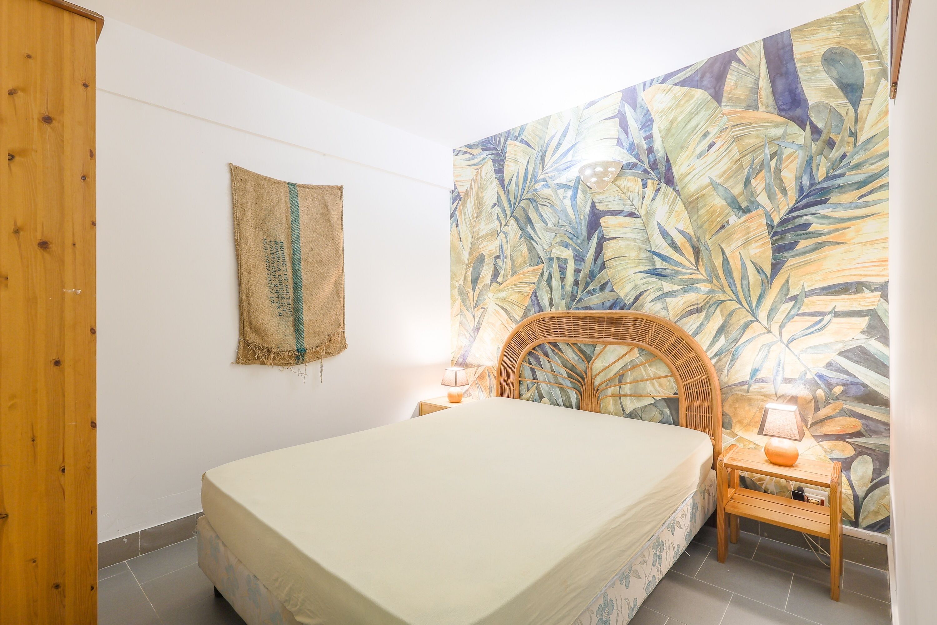 Een slaapkamer met een bed, een rieten stoel, een nachtkastje met een lamp en een muur met een tropisch bladmotief.
