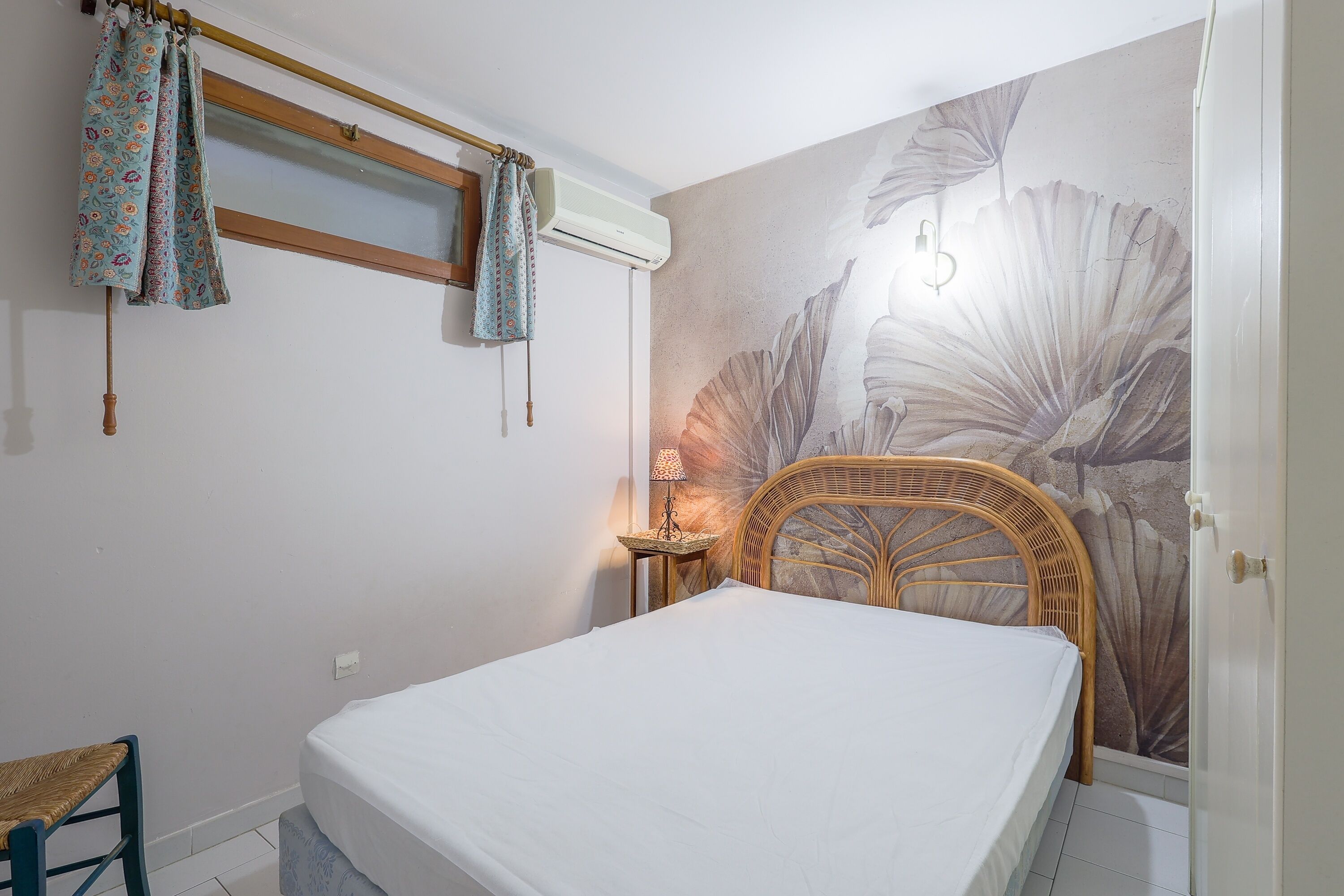 Een slaapkamer met een bed, een rieten stoel, een klein tafeltje met een lamp, een raam met gordijnen en een muurschildering.