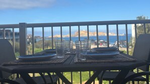 Une table dressée pour deux personnes, avec des verres et des assiettes, offrant une vue panoramique sur des collines et un plan d’eau.