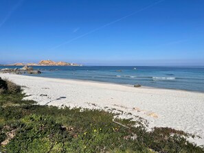En sandstrand med klart, blått vatten, klippiga utskjutande partier och ett avlägset kullområde.