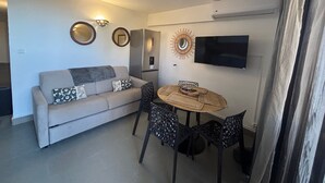 A compact living space with a sofa, a dining table, and chairs. A flat-screen TV is mounted on the wall. - L'Isula - Front de mer -  Appt pour 5 climatisé avec piscine (L'Île-Rousse)