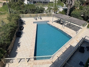 Uma piscina retangular com um deck revestido, cercada por uma cerca branca e protegida por uma pérgula.