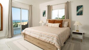 A bedroom with a large bed, two bedside tables with lamps, a view of the sea through a sliding door, and a painting on the wall.