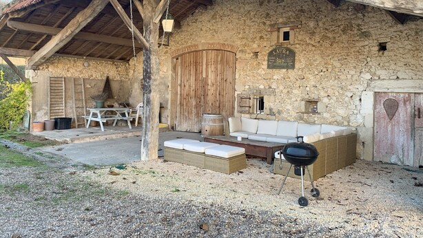 A rustic stone building with a wooden porch, a bench, a table, and a grill.