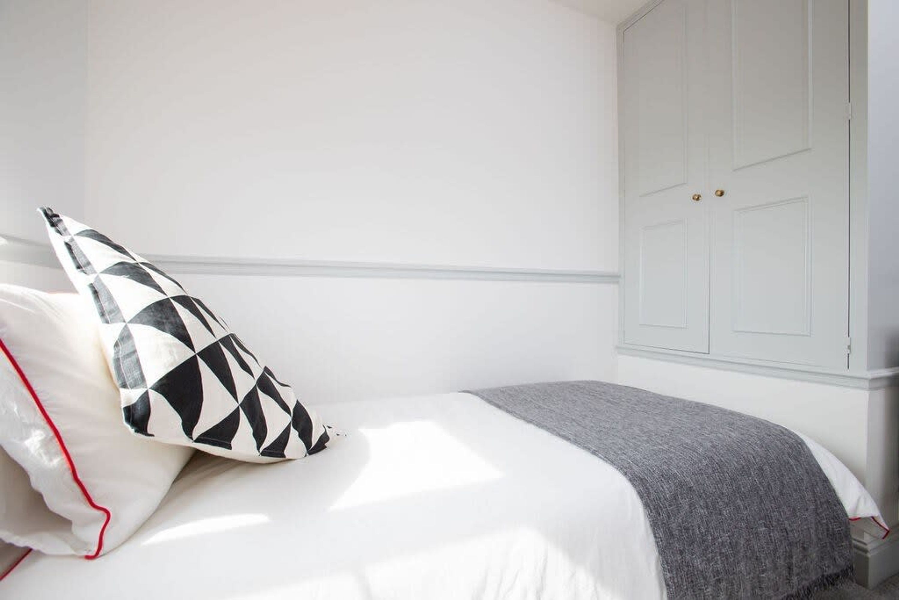 A bedroom with a bed, a white wall, a grey blanket, and a white door with two handles.