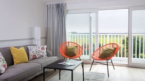A modern living room with a grey sofa, two orange chairs, a black coffee table, and large windows offering a view of greenery. - Bright Modern Selsey Seafront Home (Chichester)