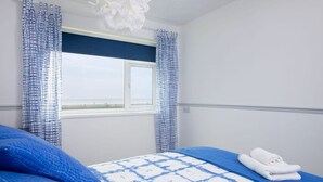 A bedroom with a bed, blue bedding, a window, and curtains. - Bright Modern Selsey Seafront Home (Chichester)