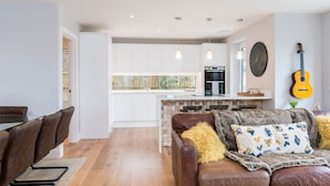 A modern living room with a brown leather sofa, a dining area with a glass table, and a kitchen with white cabinets and a built-in oven.