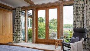 A bedroom with a large bed, a chair, a small table, and a sliding glass door leading to a garden. - Charming Cottage for 10 in Heart of South Downs (Chichester)