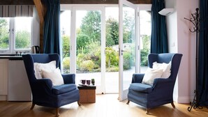 A room with two blue armchairs, a small table with a basket, and a view through a glass door to an outdoor area with greenery. - Charming Cottage for 10 in Heart of South Downs (Chichester)