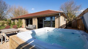 A hot tub with a wooden deck, a covered patio area, and a house with large windows and a red roof.