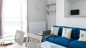 A modern living room with a blue sofa, white chairs, a wooden table, and a small white coffee table. There is a clock on the wall and a TV mounted on the wall.