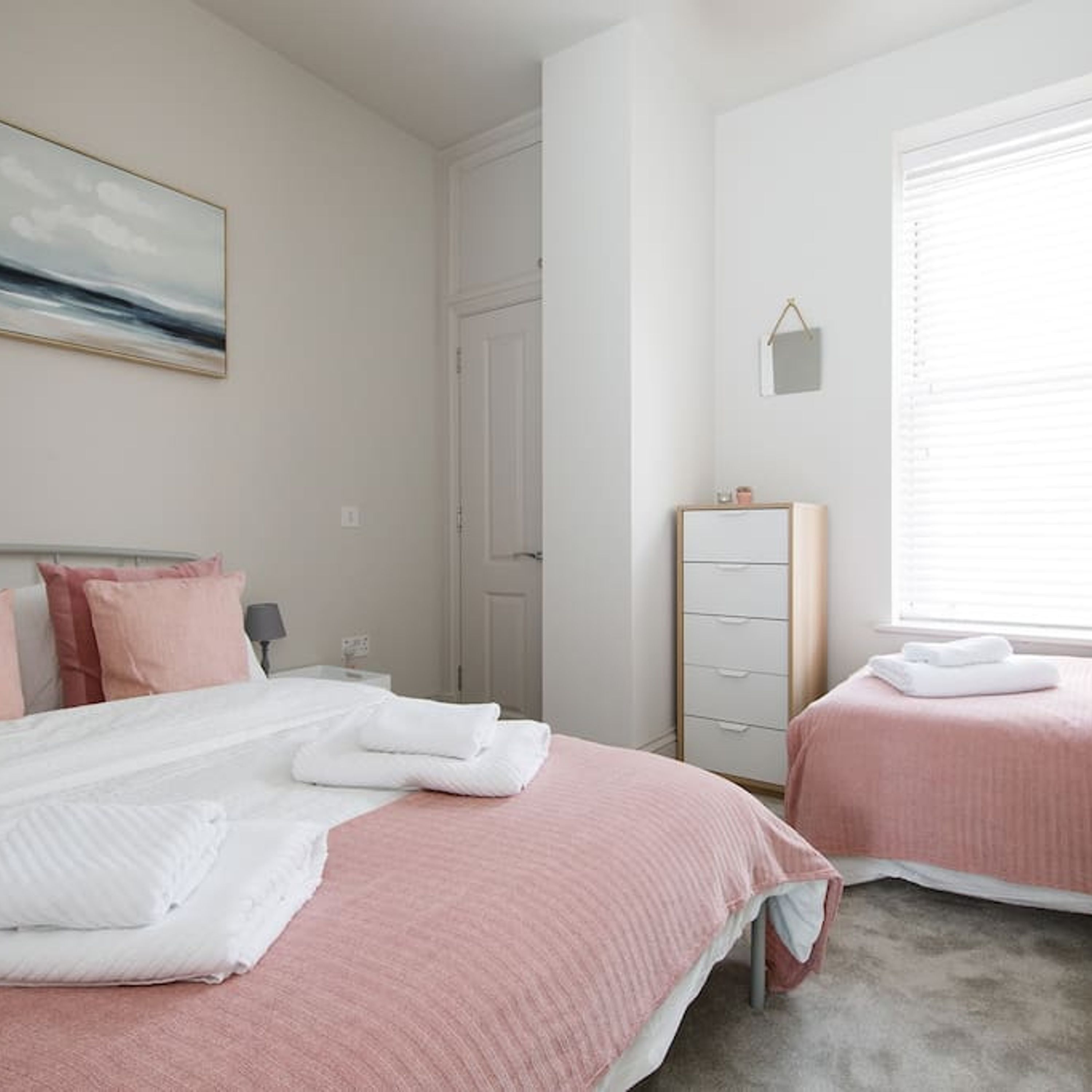 A bedroom with two beds, a dresser, a painting on the wall, and a window with blinds.