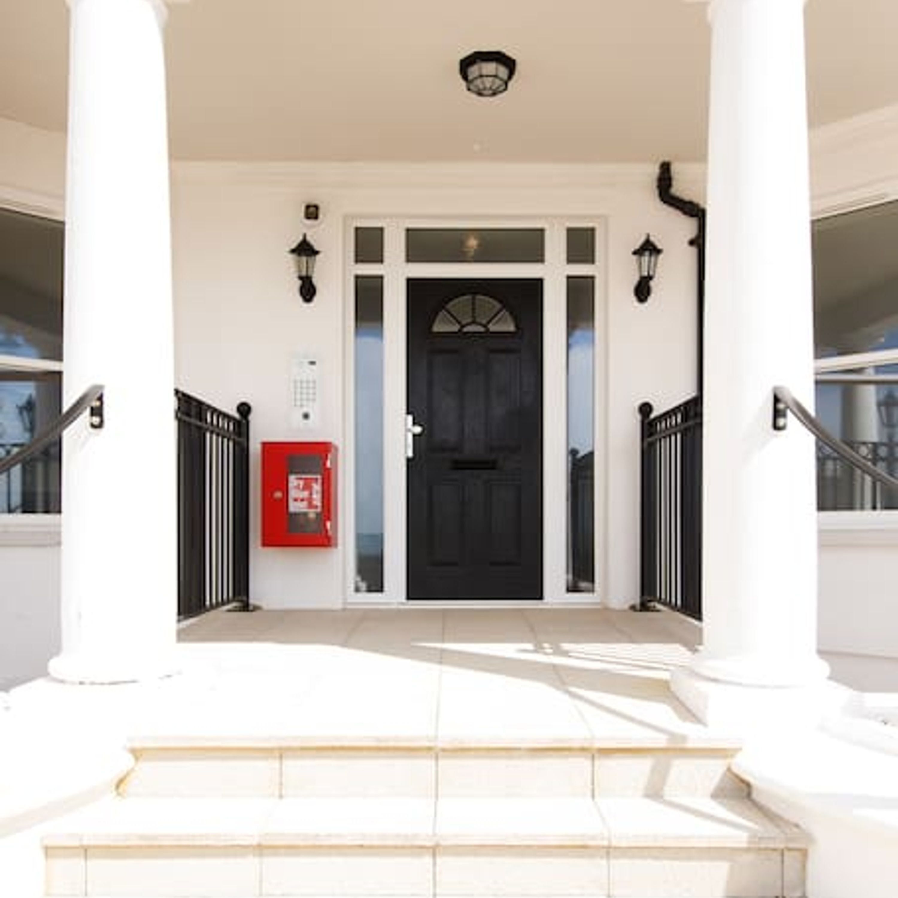 A white building with a black door, a red fire alarm, and black railings.