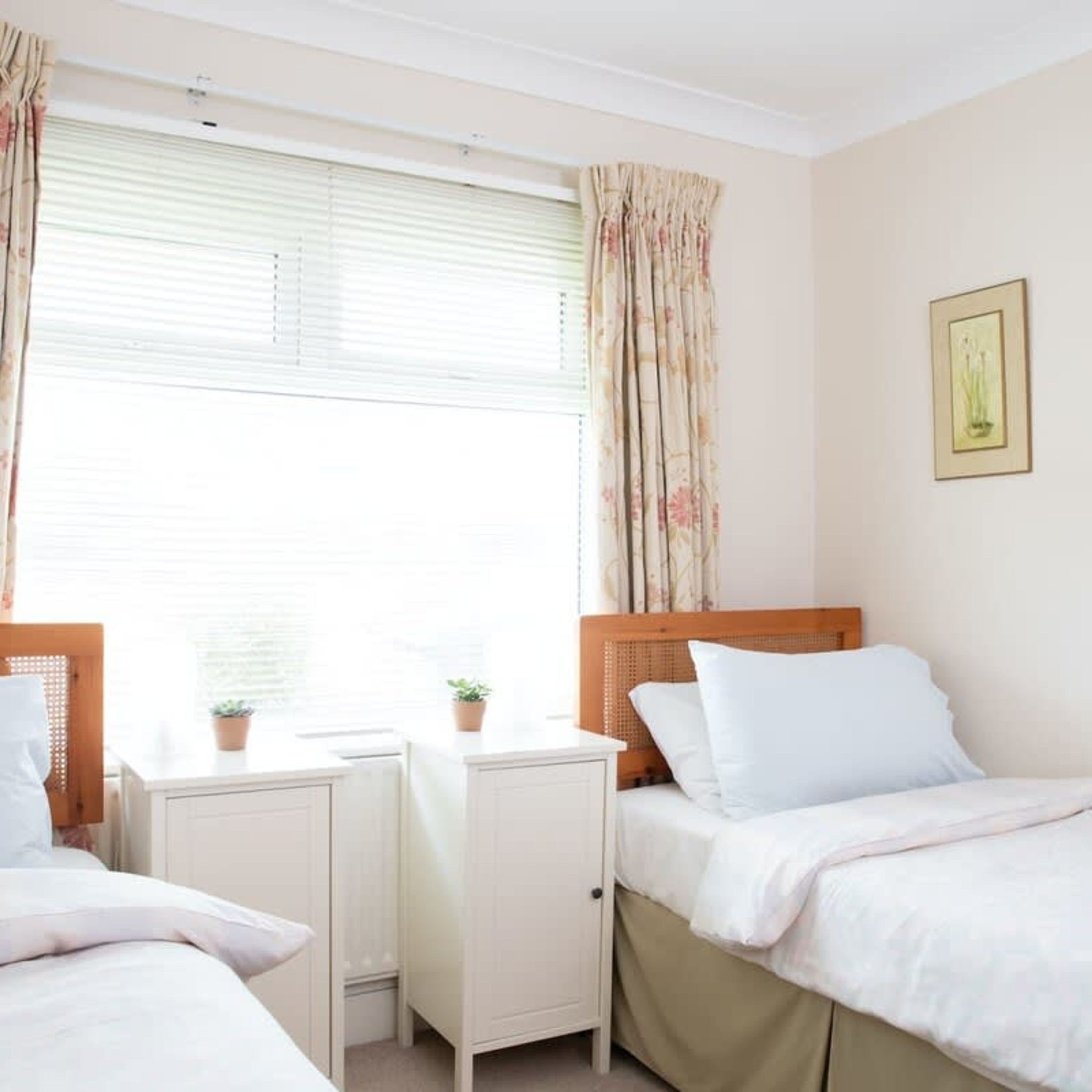 A bedroom with two beds, a nightstand, a window with curtains, and a framed picture on the wall.