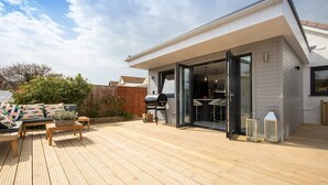 A modern house with a wooden deck, a barbecue, and a glass door leading to an outdoor dining area. - Seaside Serenity 3-bed Deck Garden Near Shore (Chichester)