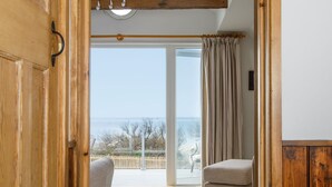 Une pièce avec une porte en bois, une vue sur la mer par une porte vitrée, une petite chaise et un ventilateur de plafond.