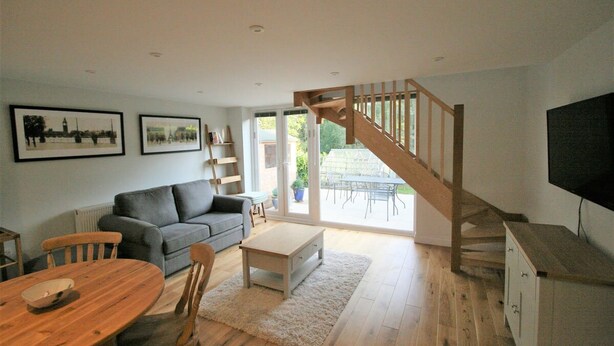 A living room with a wooden staircase, a grey sofa, a wooden dining table, and a TV on a cabinet. - Countryside Annexe Near Goodwood (Chichester)