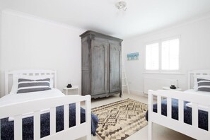 Un dormitorio con dos camas blancas, un armario gris, una ventana y una alfombra estampada.