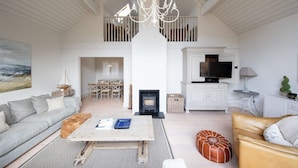 A spacious living room with a high ceiling, a fireplace, a television, and a dining area visible through an open door. - Superior House for 6 Near Beach w Parking Garden (Chichester)