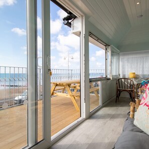 Amplia habitación con grandes ventanales que dan a una playa, una terraza de madera y un comedor con mesa y sillas.