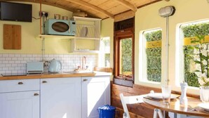 A kitchen with white cabinets, a wooden countertop, a blue trash bin, and a window with a view of greenery. - Unique Comfy Remodelled Railway Carriage (Chichester)