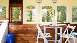 A wooden cabin with a dining area featuring a round table and white chairs, a window with a view of greenery, and a blue trash bin. - Unique Comfy Remodelled Railway Carriage (Chichester)