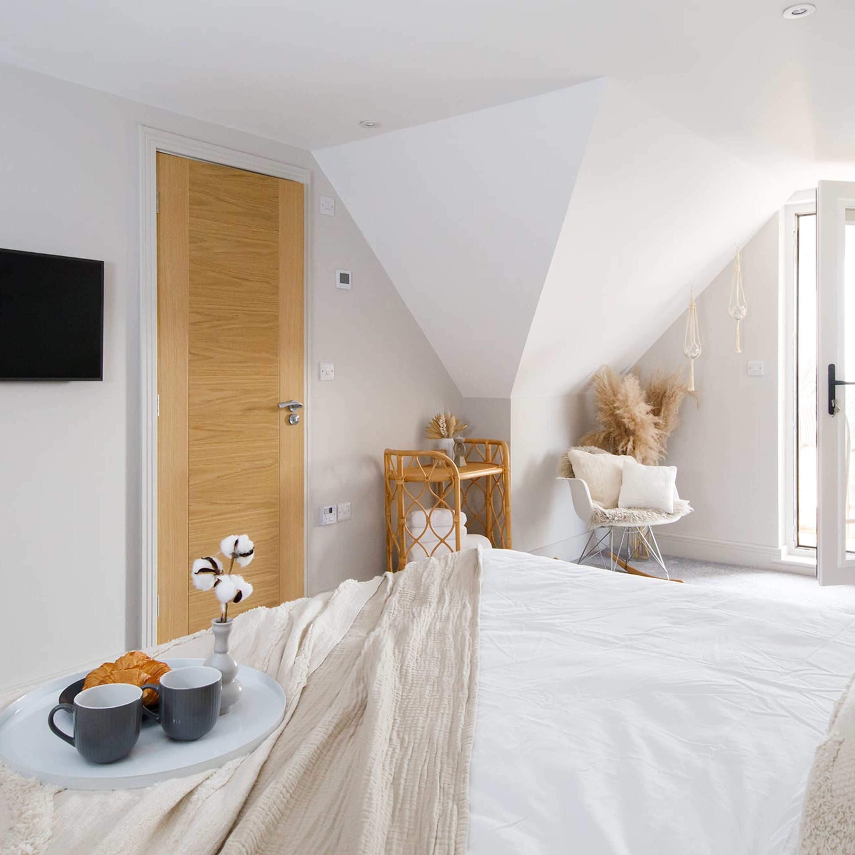 Een moderne slaapkamer met een schuine plafond, een houten deur, een bed met witte beddengoed, een klein rond tafeltje met twee mokken en een croissant, en een stoel met een witte kussen.