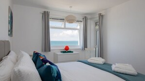 A bedroom with a bed, pillows, a bedside table with a lamp, a window with curtains, and a view of the sea.