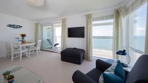 A modern living room with a glass table, white chairs, a black ottoman, a flat-screen TV, and a view of the sea through large windows.