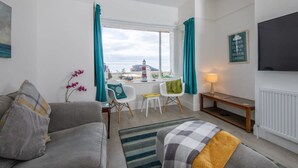 A modern living room with a sofa, a coffee table, and a TV. There is a window with curtains, a small table with chairs, and a lamp. - Absolute Beachfront With Decked Garden Oasis and Views (Bognor Regis)