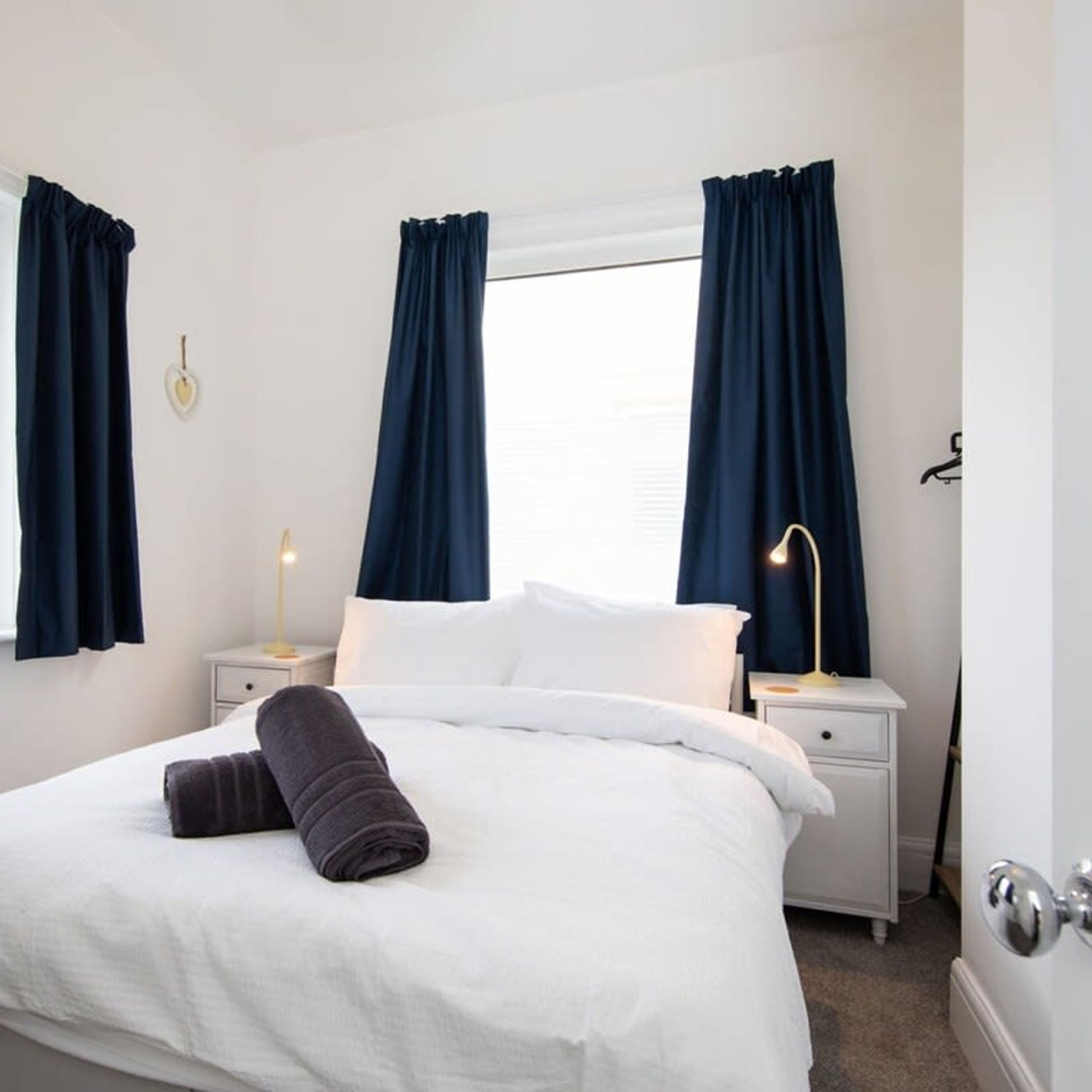 A neatly made bed with white linens and a rolled-up dark towel, two bedside tables with lamps, and a window with blue curtains.