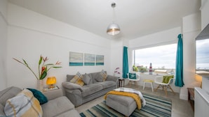 A modern living room with a grey sofa, ottoman, and a striped rug. There is a dining area with a table and chairs, and a view of the sea through large windows. - Absolute Beachfront With Decked Garden Oasis and Views (Bognor Regis)
